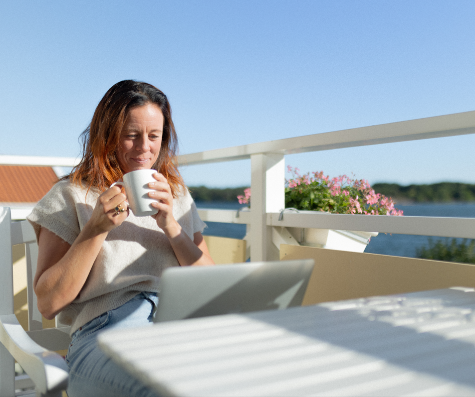 Kvinna som njuter av sitt kaffe på balkongen, med en laptop framför sig.