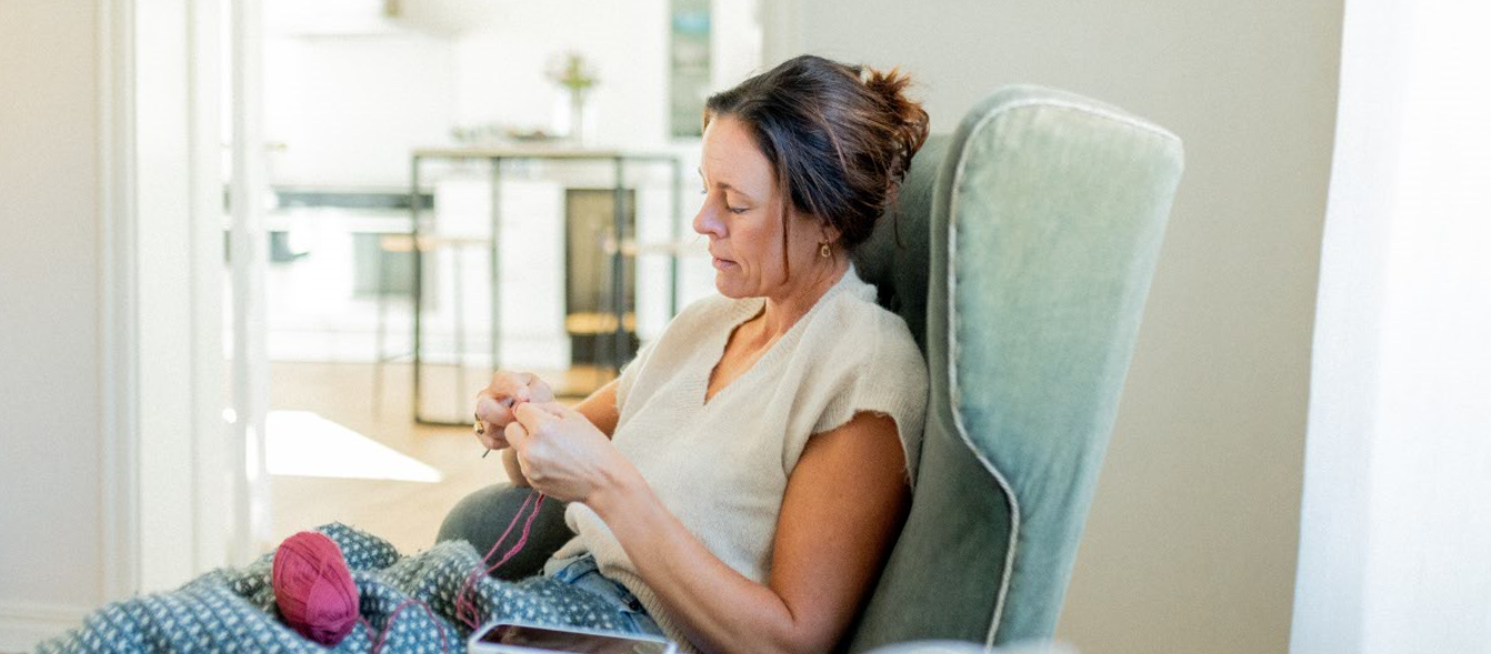 Kvinna som sitter och stickar med mobiltelefonen på laddning.