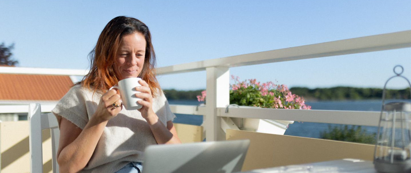 Kvinna dricker kaffe framför sin laptop på sin balkong.